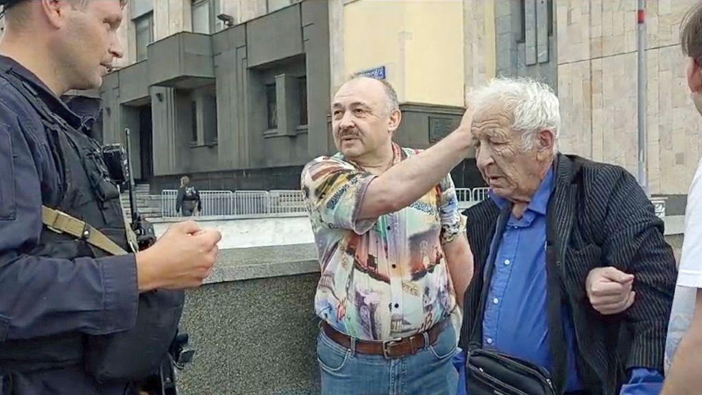 An elderly man, with grey hair, a suit jacket and blue shirt, is frowning, while two men put their arms through his, holding him. The man on the left of him has a boldly-patterned shirt and moustache, and is talking while gestring to the elderly man. A police officer in navy uniform is looking at both of them.