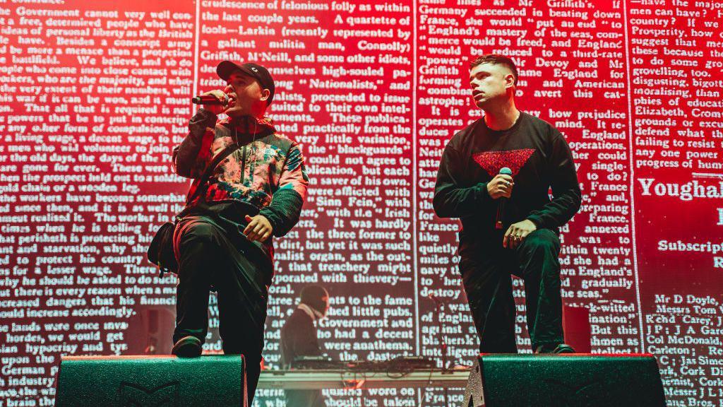 Two men, one weaing a jacket and a baseball cap, and the other wearing a jumper, stand in the foreground. Behind them, a third man stands wearing a tri-colour balaclava. The two men at the front are holding microphones. The three men are on stage, and behind them is a red screen with white writing on it. 