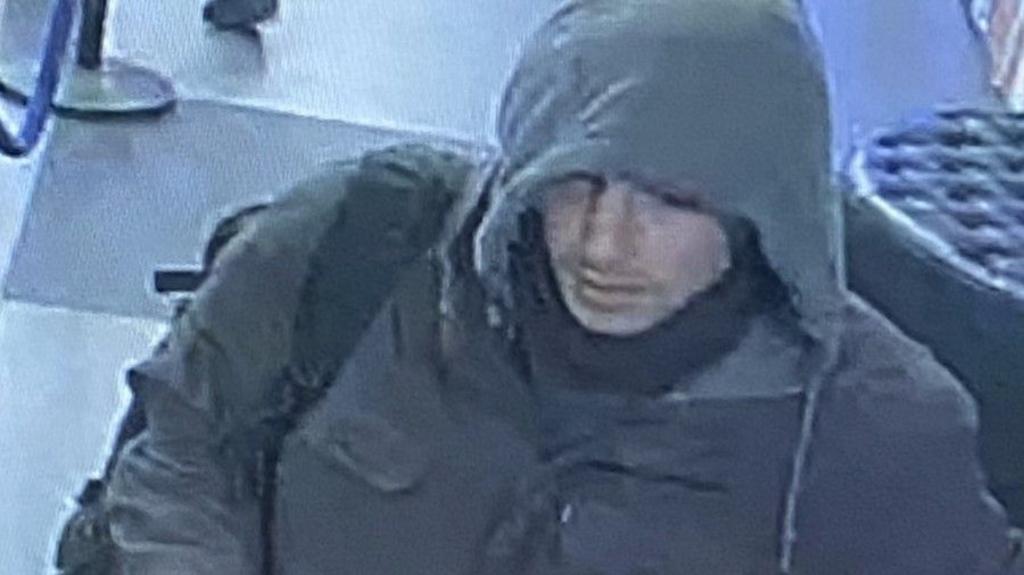 A man in a hooded jumper wearing a black jacket is show on a cctv image.