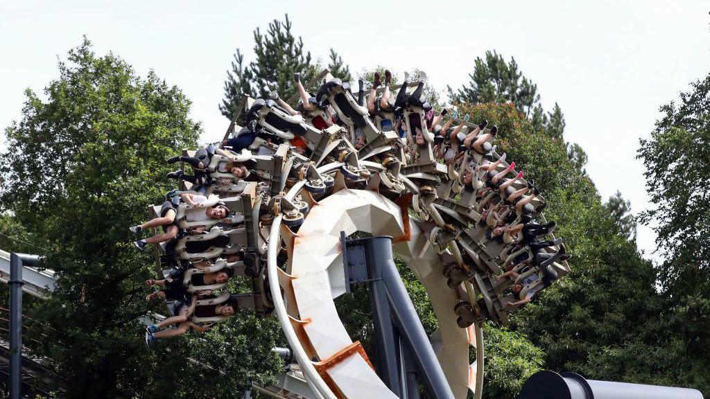The Nemesis rollercoaster - showing a section of its track where a carriage of riders are in the middle of a rotation, with some riders off to the side of the ride, and some upside down - some trees are visible in the background