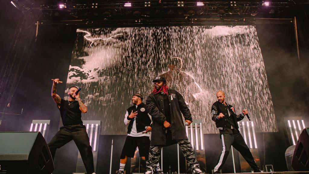 The four members of JLS on stage singing. There is a screen behind them playing a music video with rain pouring down. They are in black clothes and have handheld black microphones