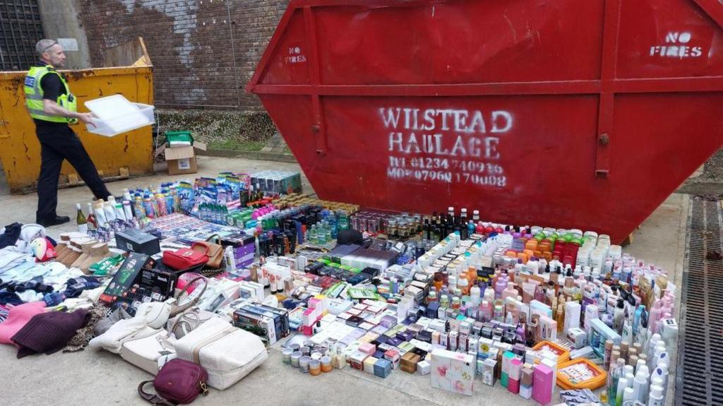 A red waste skip with the words Wilstead haulage in white paint stands on concrete, with hundreds of goods laid on the ground in front of it, including toiletries, household products, handbags, and other bottles and products which are unidentifiable. A police officer stands beside it.