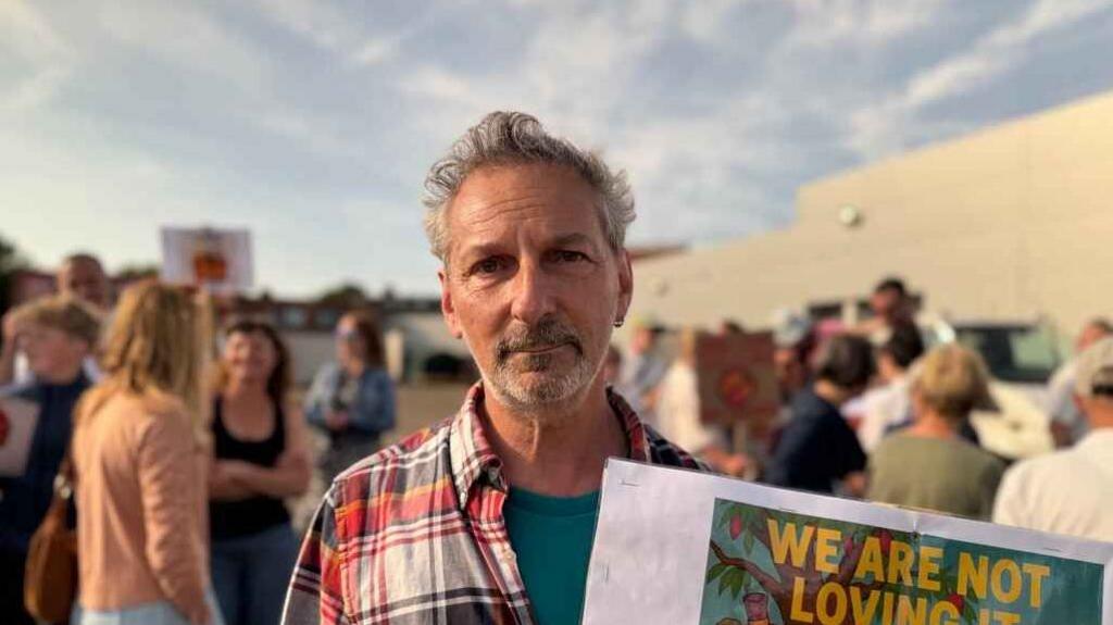 Charles Rickards wearing a striped shirt and holding a placard which reads "we are not loving it". he has a crowd behind him also holding signs