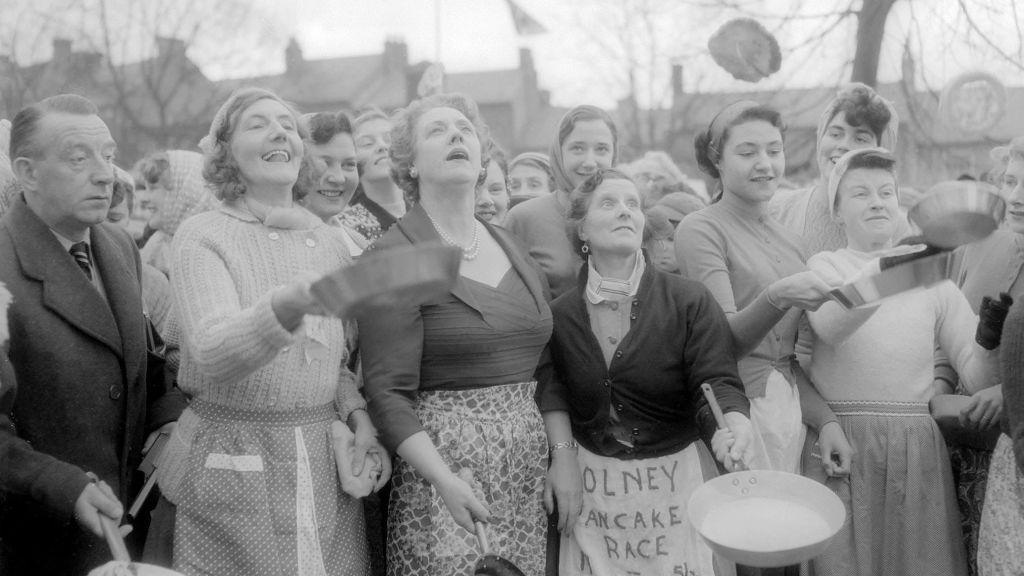 
A black and white photo from the 1950s of women and one man holding frying pans and flipping pancakes. 