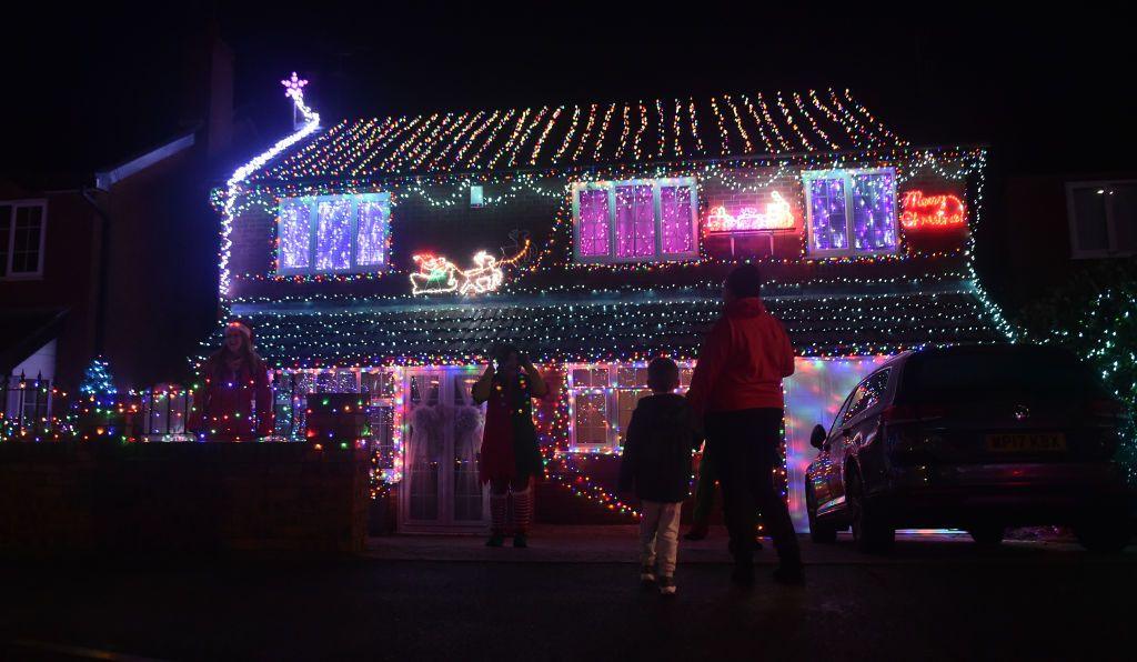 A house is covered in Christmas lights which are lit up in the dark as people look at it from the pavement. 