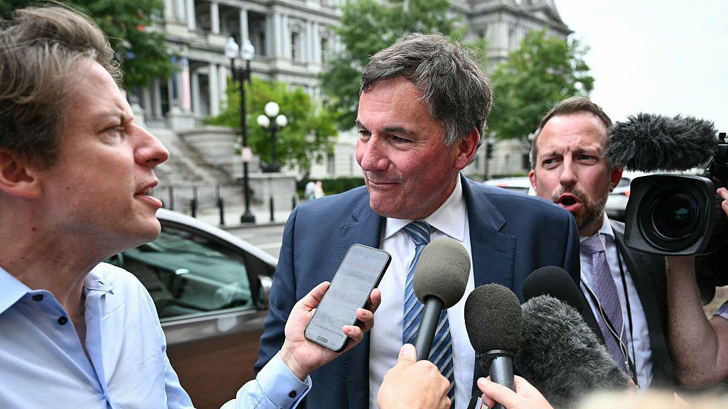 LeBlanc is pictured surrounded by reporters on a side walk in Washington, DC. He has short black and grey hair and is listening to a reporter asking him a question. The reporter holds up an iPhone to LeBlanc's face to record him. Others hold up microphones to his face. A TV camera is on his right. He is wearing a navy blue suit and a striped blue tie.