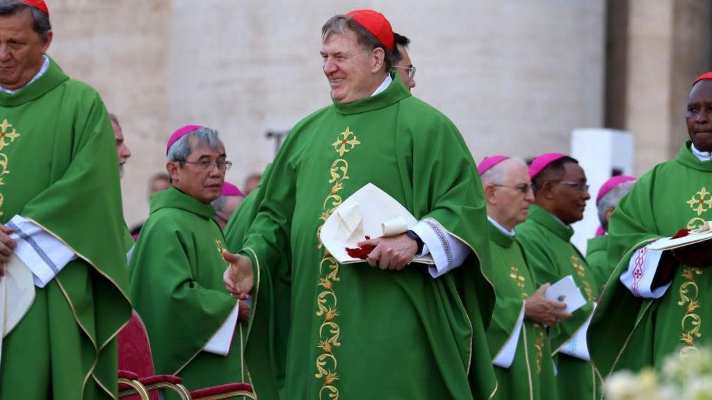 Cardinal Joseph Tobin wears a large green cloak and a red skullcap as he grins at the Vatican in 2022