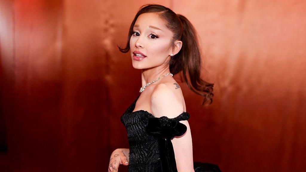 Ariana grande with her signature ponytail wearing a black dress and looking over her shoulder act the camera