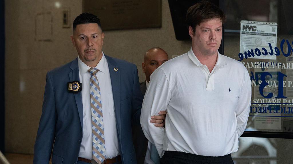 William Duplessie wearing a white dress shirt is being led by police detective who is gripping his arm. 