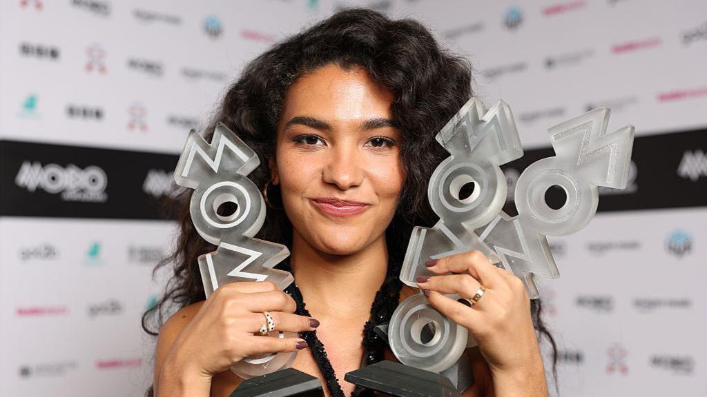 Olivia Dean facing the camera holding three Mobo trophies