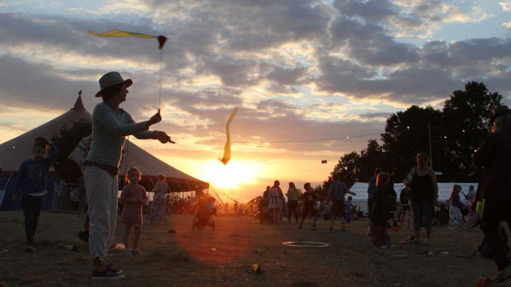 A man wearing a hat waves two sticks in the air. The sticks have yellow ribbons on them. In the background, the sun is setting and there is a large festival tent. A number of punters can be seen walking around, although most of them are silhouettes.