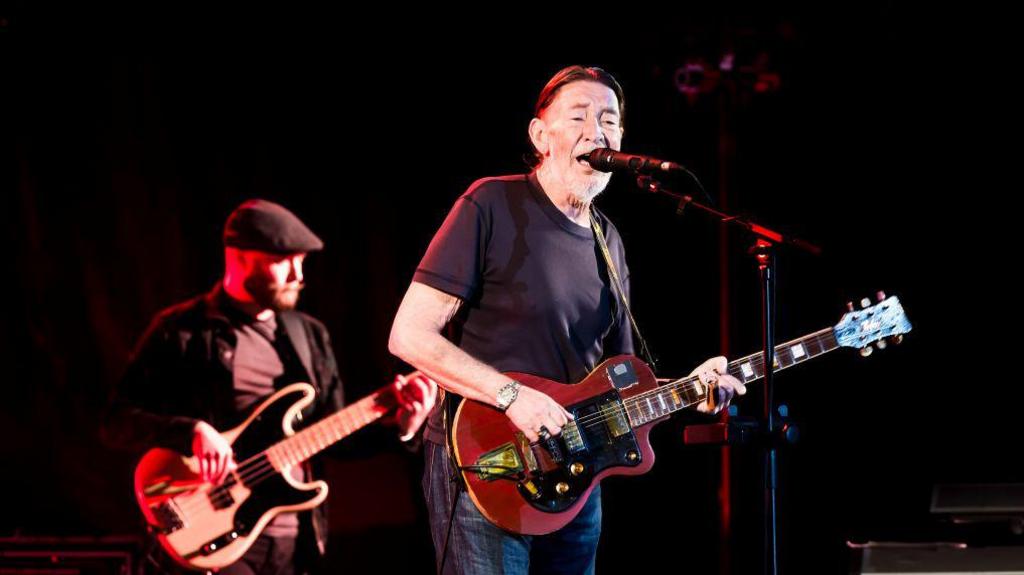 British singer Chris Rea stands at the mic, holding a guitar. He is in the middle of singing in this picture. He has black hair slicked back and white stubble. .He wears a black t-shirt.  The picture was taken during a concert at the Tempodrom on October 30, 2017 in Berlin, Germany.