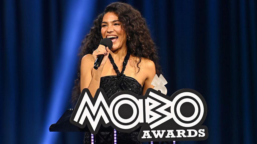 Olivia Dean smiling into a microphone standing in front of a podium that says 'Mobo Awards'