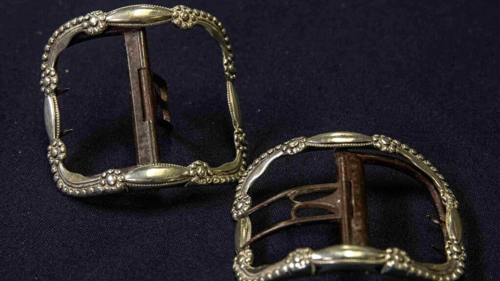 Two 19th century shoe buckles are displayed on a felt cloth and photographed from above. They are elaborately decorated, and a dim gold in colour. They have a ribbon pattern around the outside and a rusty metal fixing in the middle of both. 