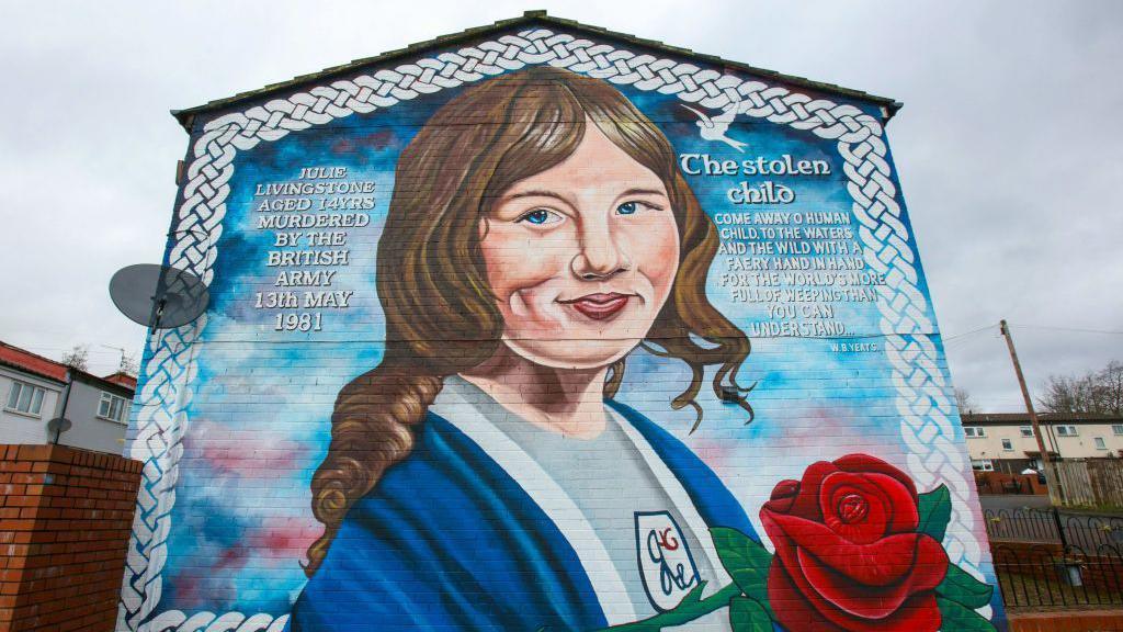 A mural on a gable wall. This shows a girl with brown hair holding a rose, she is wearing a blue shawl over a grey top. The words 'JULIE LIVINGSTONE AGED 14YRS MURDERED BY THE BRITISH ARMY 13TH MAY 1981'  is written on the left. On the right the words 'The stolen child - COME AWAY O HUMAN CHILD TO THE WATERS AND THE WILD WITH A FAERY HAND FOR THE WORLDS MORE FULL OF WEEPING THAN YOU CAN UNDERSTAND - WB Yeats' .