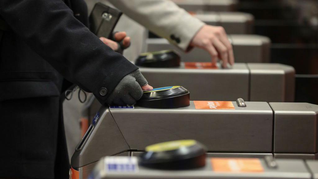 A close-up file image of a person wearing a dark coat and grey fingerless gloves tapping their blue and white Oyster card against the round yellow reader at a ticket barrier in a station building. A person wearing a light coat can be seen behind them reaching for a ticket being fed out of the reader at another barrier further along.