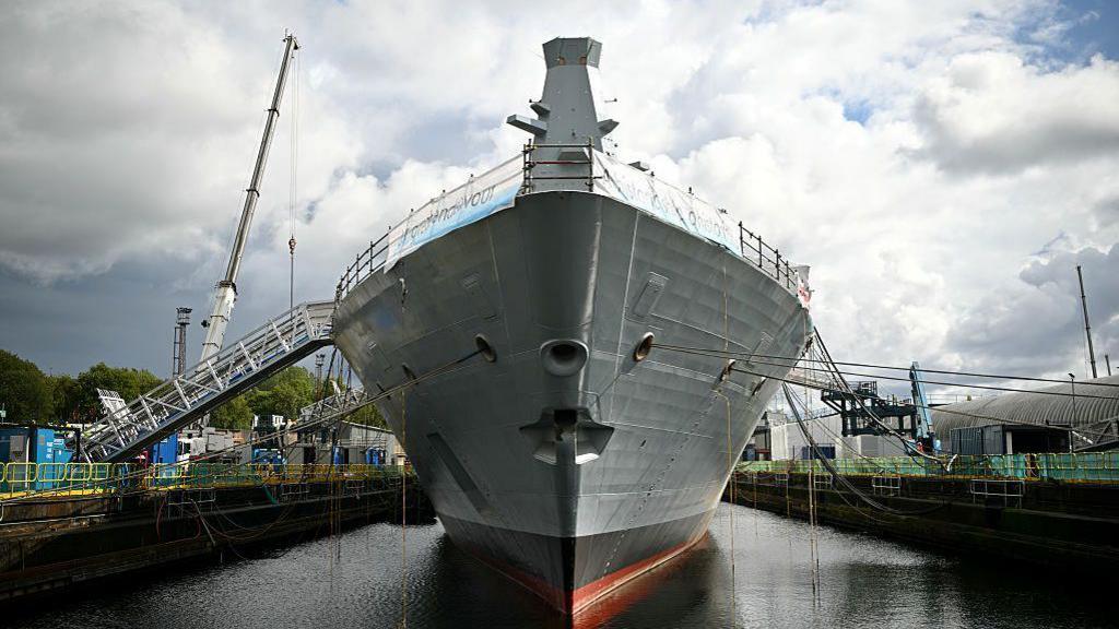 A large warship, seen front on in a dock with a crane in the background. 