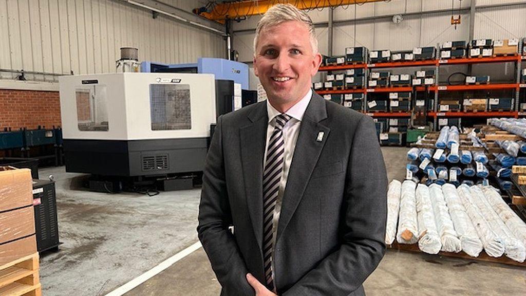 William Cook smiles at the camera. He is clean shaven with short neat grey hair and is wearing a dark suit, white shirt and grey and white striped tie. Behind him are various pieces of factory equipment, racks with boxes on and piles of metal pipes.