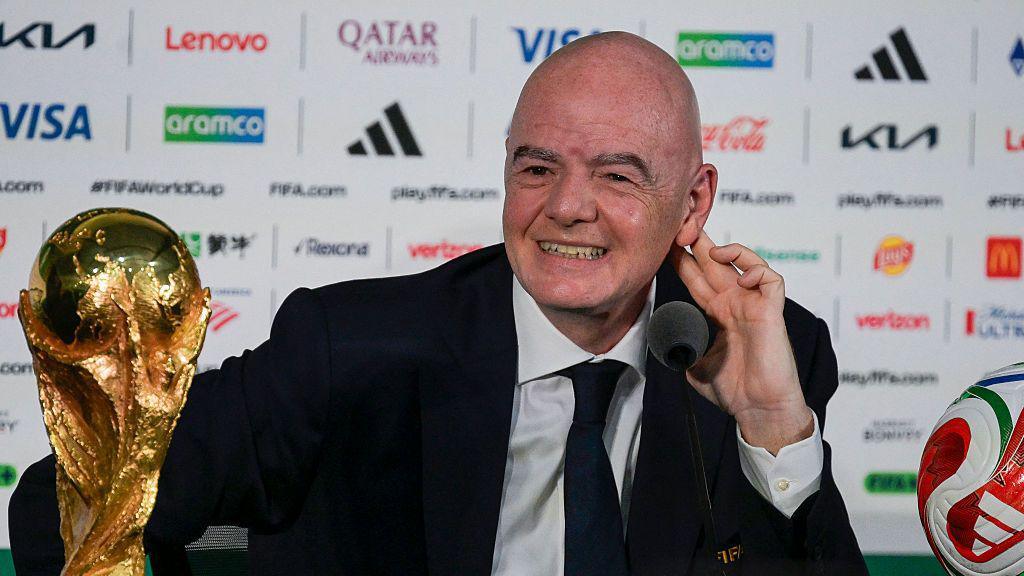 FIFA president Gianni Infantino, in dark suit and tie and white shirt, speaks next to the World Cup trophy and a football during a news conference on the eve of the opening match of the 2026 World Cup