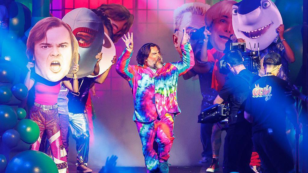 Jack Black wears a tie dye suit and is gesturing to the audience. Behind him are people holding up large cardboard cutouts of the characters he has voiced over the years