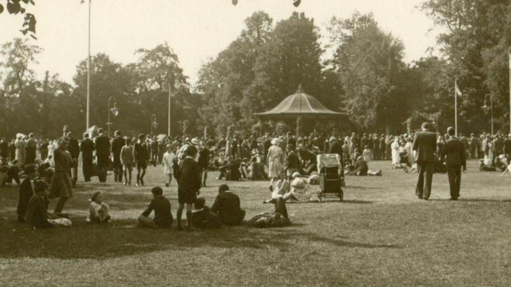 A black and white photo of people in a park. There is a bandstand in the distance with a conical roof. Some people are sitting on the grass while others are walking around. There is a large group of people near the bandstand. There are trees in the background.