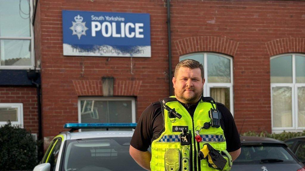 A police man in a yellow vest stands with his arms behind his back in front of a red brick police station