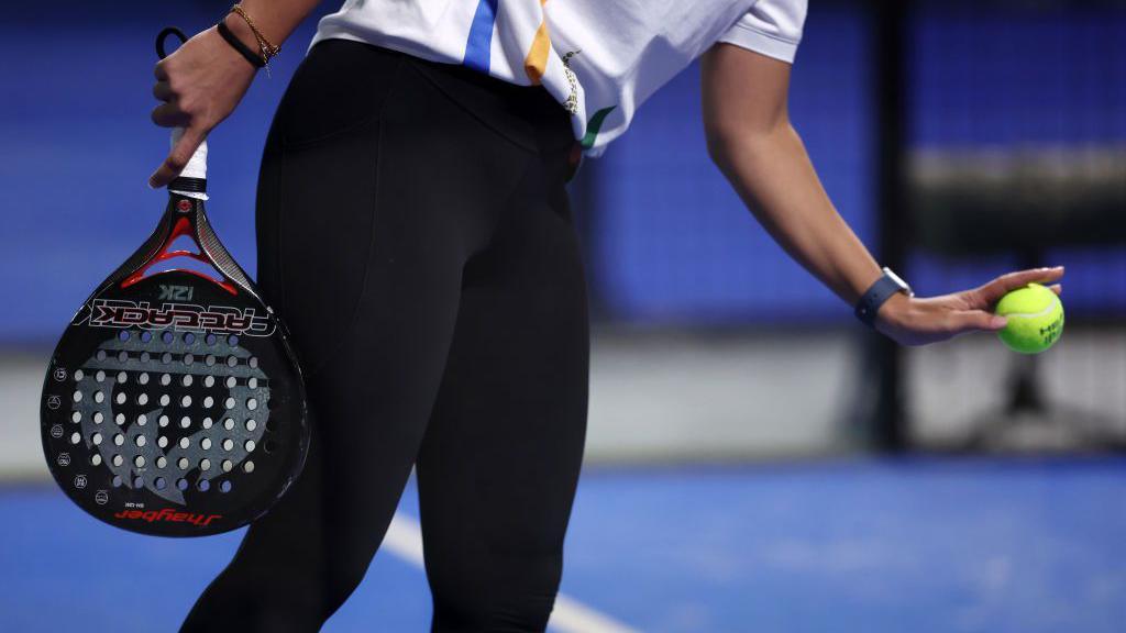 A woman wearing a white patterned T-shirt and black leggings prepares to serve a light green tennis ball with a black padel racquet. It has a short handle and solid surface with air holes through it, unlike a traditional stringed racquet.