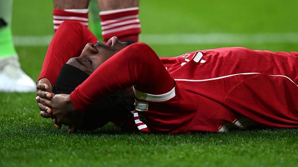 Liverpool's Jeremie Frimpong lies on his back on the pitch, injured, with his hands on his head. He is wearing Liverpool's red home kit.
