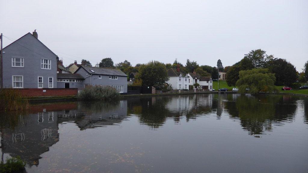 A large pond with houses running parallel to it. There are also large trees and cars parked in the background. The picture has been taken on a grey day.