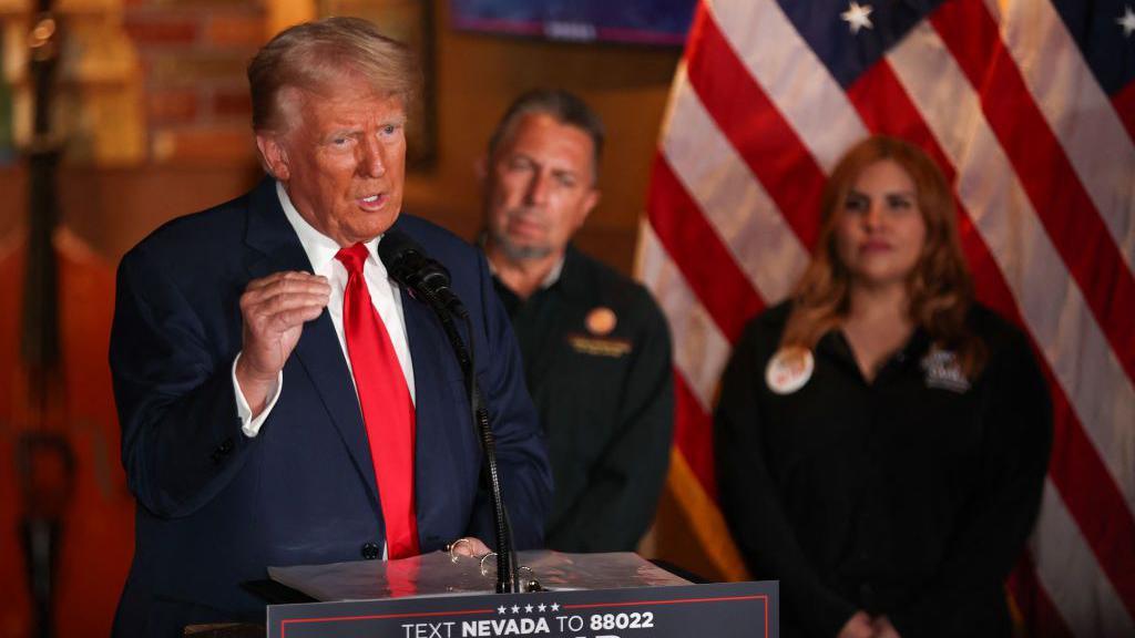 Donald Trump alongside Las Vegas restaurant owner Javier Barajas and another person. There is a US flag in the background. Trump is stood at the front, speaking into a microphone