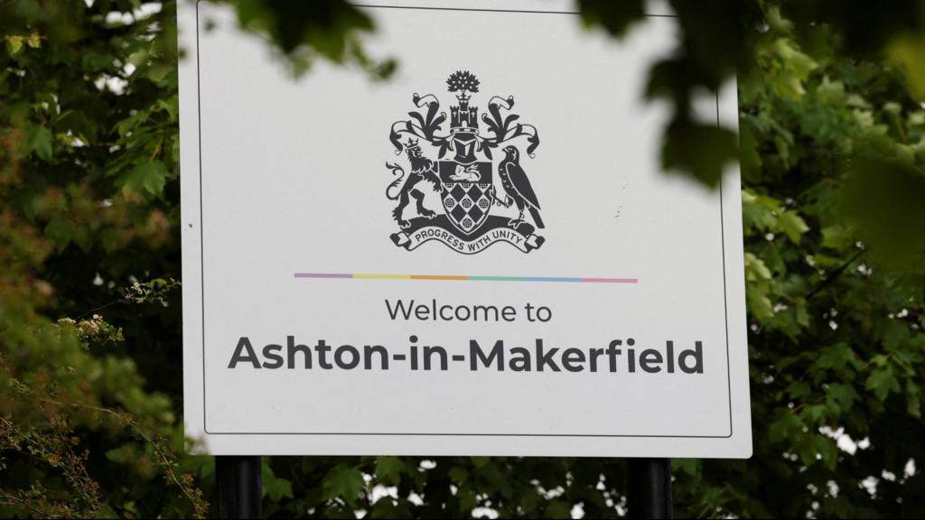 A white sign with black writing reading 'Welcome to Ashton-in-Makerfield'