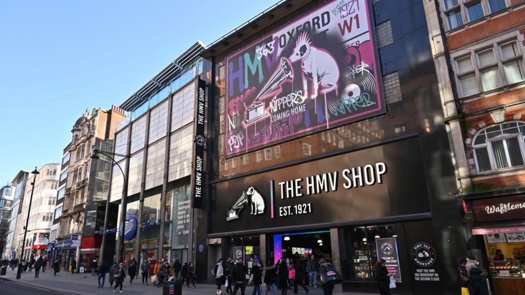A large music retail shop, the HMV shop. It has a logo of a white dog listening to a gramophone. There are lots of people walking across the shop on a busy street in London.