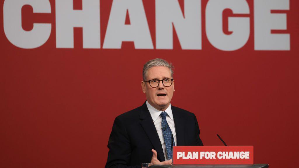 Sir Keir Starmer giving a speech on a podium with the words "Plan for Change" on the lectern (red background and white font). The slogan is also the background behind him - only change is visible.