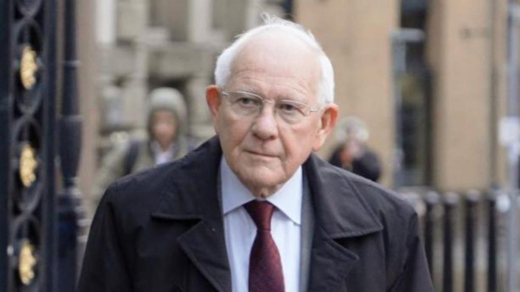 Sir Duncan Nichol wearing a blue shirt, burgundy tie and black coat walking past some iron railings