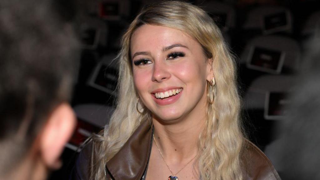 Haliey Welch shown in a close up photograph at an event in November, wearing a brown leather jacket and smiling.