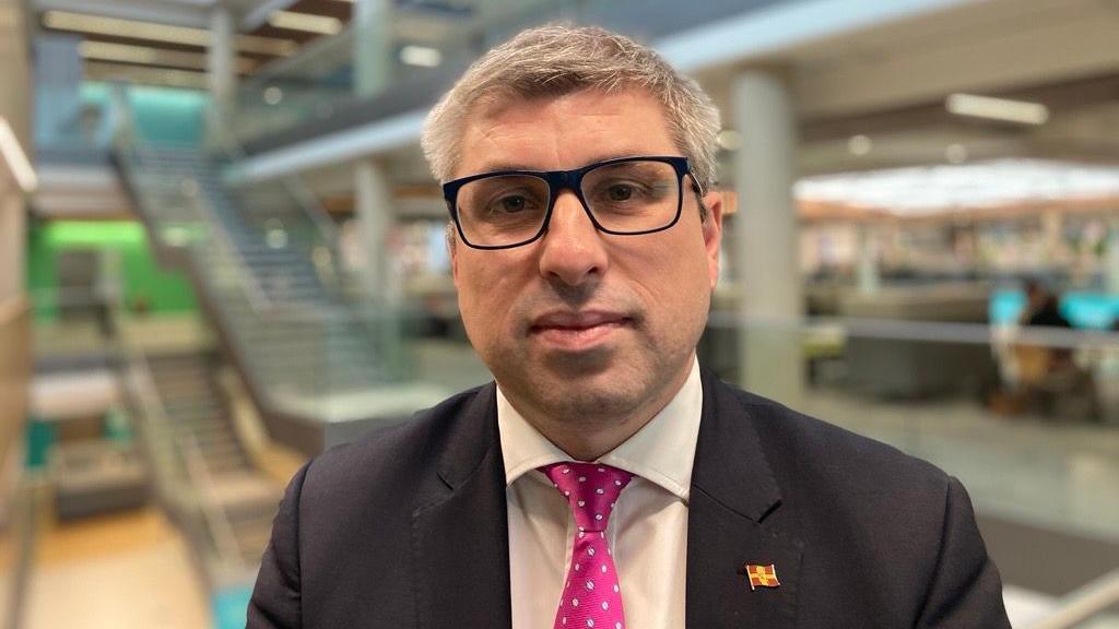 Mark Arnull, who has short hair and black framed glasses. He is wearing a dark jacket over a white shirt and pink tie. Behind him is an office setting.