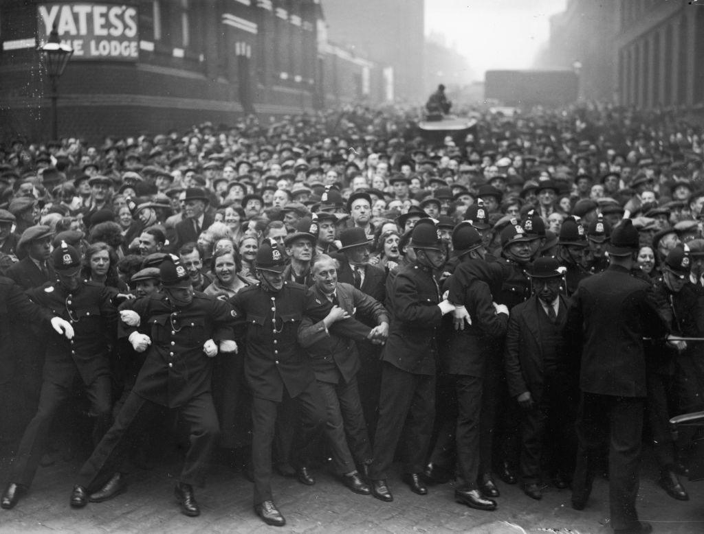 A row of police officers hold back a crowd of hundreds of people.