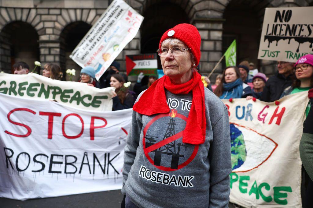Protesters outside Edinburgh's Court of Session with white banners saying "stop Rosebank". In the foreground is a woman with glasses, a red hat and scarf, and a grey jumper with an anti-Rosebank slogan