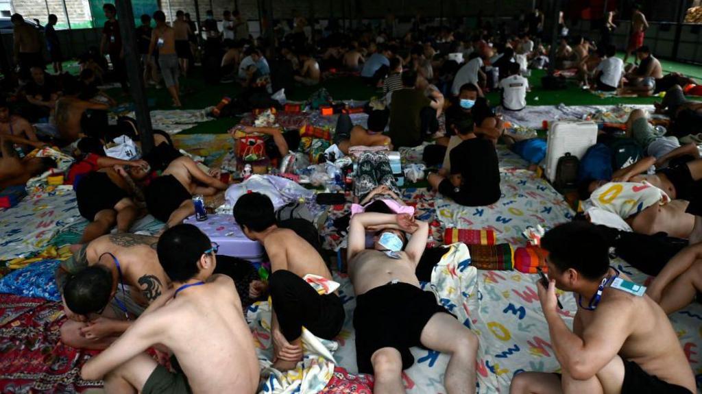 A roomful of men, some lying down on mattreses, others sitting up or standing, in a makeshift camp. Many of them are shirtless and in shorts. 