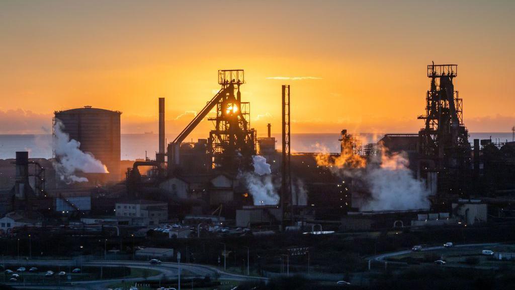 The steelworks in Port Talbot at sunset. White steam rises from the chimneys. 