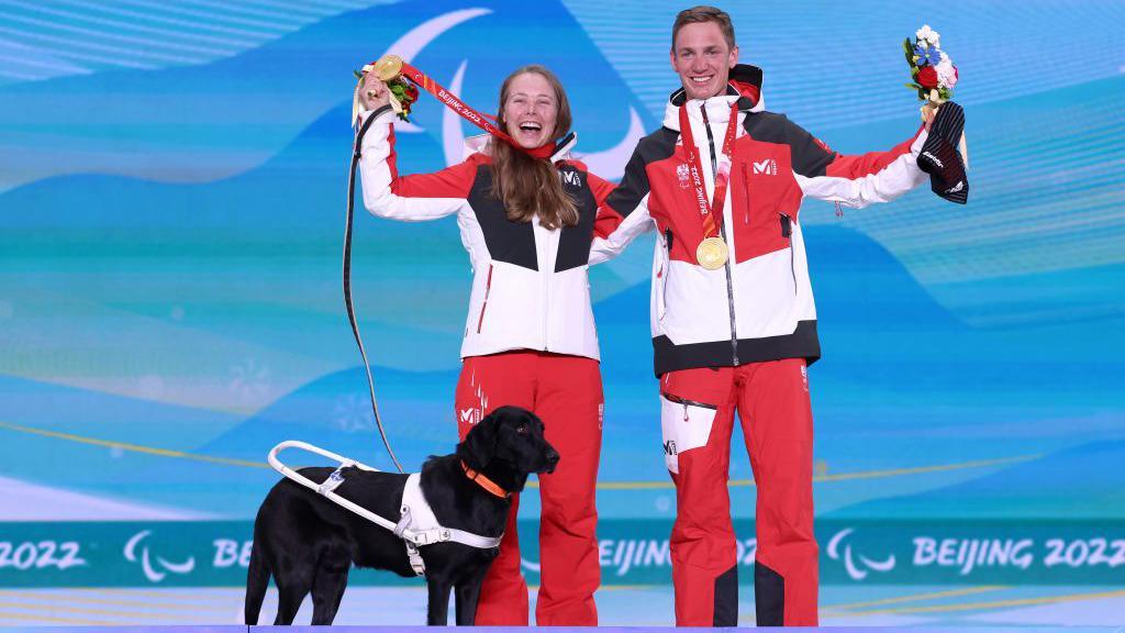 Austria's Carina Edlinger smiles and celebrates holding her gold medal in the air alongside her guide Lorenz Lampl and her assistance dog, a black labrador called Riley, on the podium at Beijing 2022