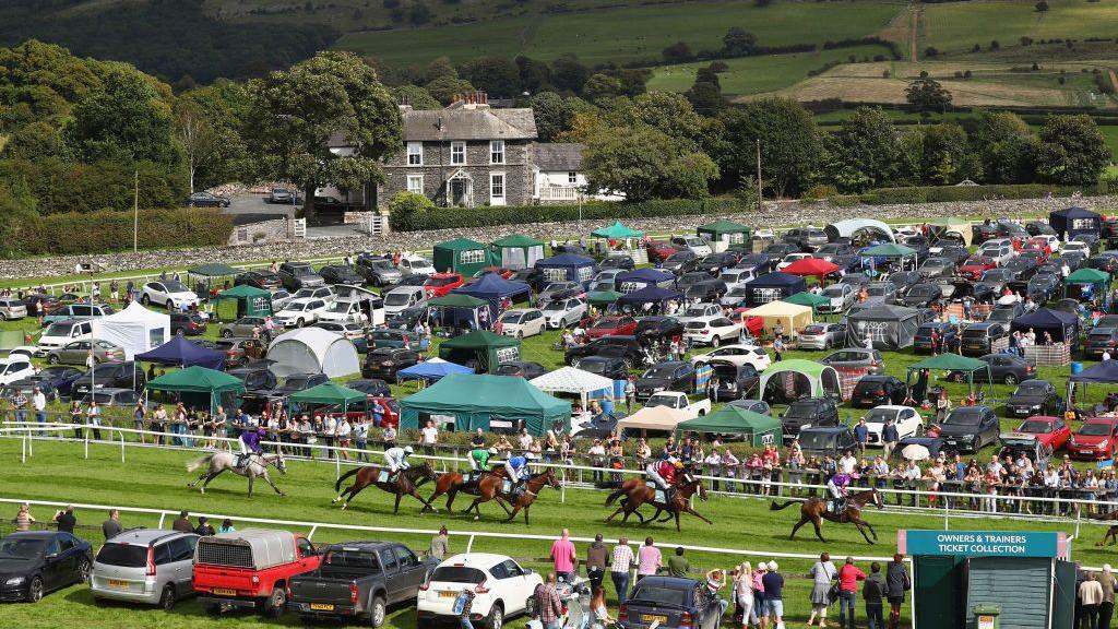 Cartmel Racecourse pictured during the Hadwins Motor Group Handicap Hurdle race in 2017. Horses are racing and the course is surrounded by cars and gazebos.