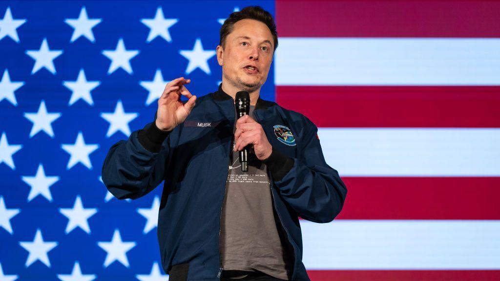 Elon Musk holds microphone and stands in front of a large American flag