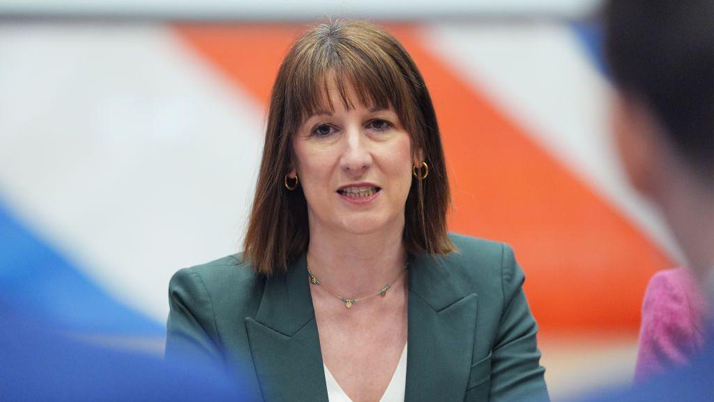 Headshot of Chancellor Rachel Reeves in a green suit with part of the Union Jack behind her