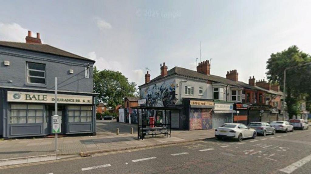 A Google street view image of King Richards Road showing a path leading to Kate Street in Leicester. There are a number of parked cars on the main road in front of businesses in the street.