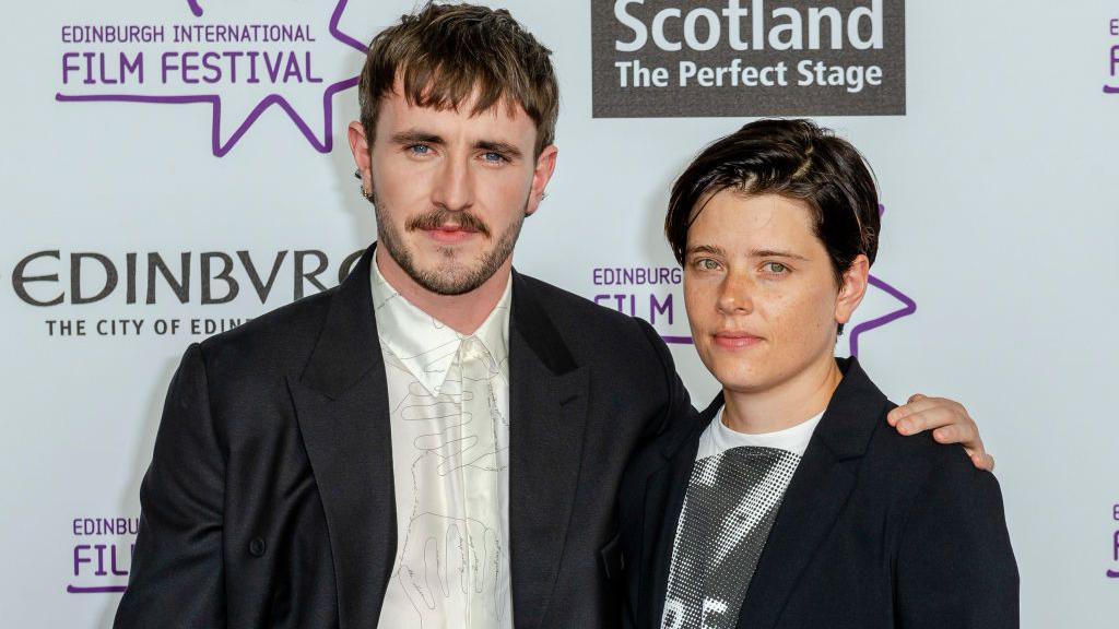 Paul Mescal and Charlotte Wells in monochrome outfits pose against a sponsor backdrop with logos of the Edinburgh Film Festival, Visit Scotland and the city of Edinburgh.