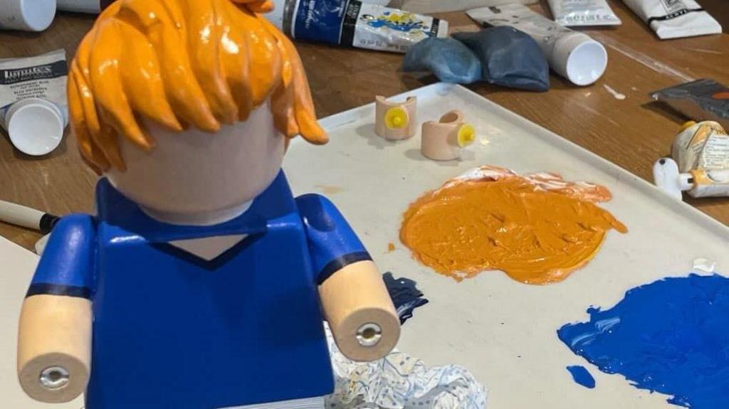 A photo of the figure before it had most of the details painted on. It has ginger coloured hair and an blue top painted on but it is missing its facial features and the hands are yet to be added. A paint pallet can be seen sitting next to it with the orange and blue colours on it.