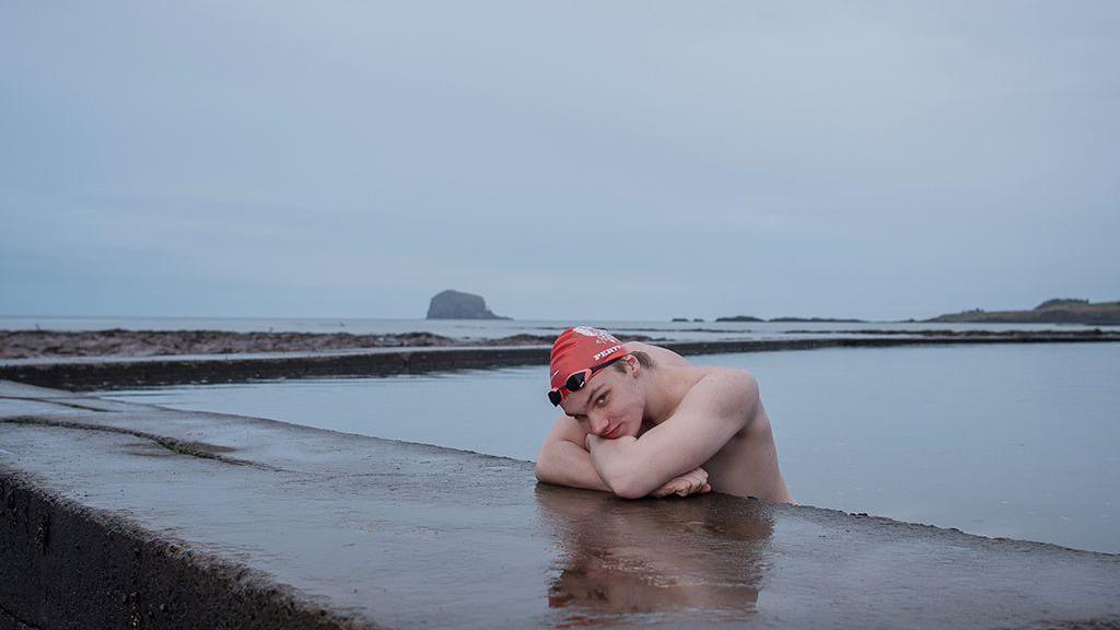 Male swimmer with red swim cap leans on concrete at side of outdoor pool, looking at camera, against grey background.