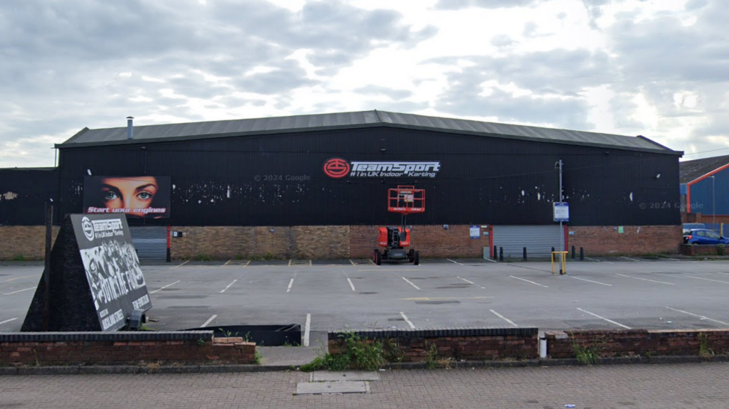 The building is black and brick with its sign on one wall in white writing. There is a car park in front of it.
