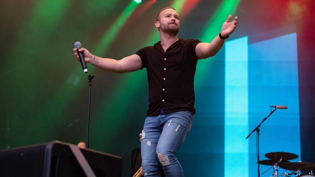 A singer with his arms open in a black shirt and jeans encourages the crowd to join in